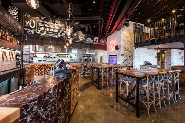 A dramatic industrial-style restaurant and bar interior with a polished dark marble bar in the foreground, exposed black ceiling services and red pipework, white-painted brick walls, chalkboard-style menu signage, timber accents, metal-framed tables, and woven bar stools. A mezzanine with black metal balustrading overlooks the dining area, while the open kitchen and wood storage are visible in the background.