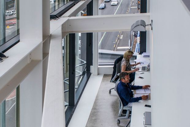 A modern commercial office interior viewed from an elevated angle, with two people working at a long white shared workstation beside floor-to-ceiling windows. The space features exposed white structural beams, dark-framed glazing, pale carpet, and views of an urban roadway outside.