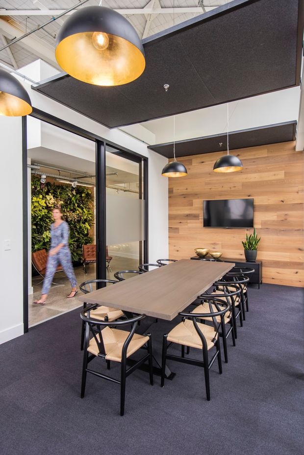 A contemporary commercial meeting room with a long timber dining table surrounded by black-framed chairs. The room features dark carpet, black acoustic ceiling panels, exposed white services and roof structure, black-framed glazed partitions, pendant lights with black exteriors and warm metallic interiors, and a timber feature wall with a mounted television and low storage unit. A planted green wall is visible through the glass partition.