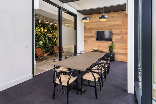 A contemporary office meeting room with a long timber meeting table surrounded by black-framed chairs. The room features dark carpet, black-framed glazed partitions, a warm timber feature wall with a mounted television, pendant lighting, and a visible planted green wall in an adjacent space.