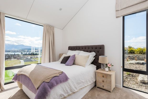 Bright, contemporary bedroom with a tufted dark upholstered bedhead, white bedding, and layered beige and lavender throws and cushions. A small timber bedside table with a lamp and flowers sits beside the bed. Full-height glazing on two sides provides expansive views of a landscaped property and distant water and hills, with neutral curtains, a roman blind, and soft carpet completing the room.