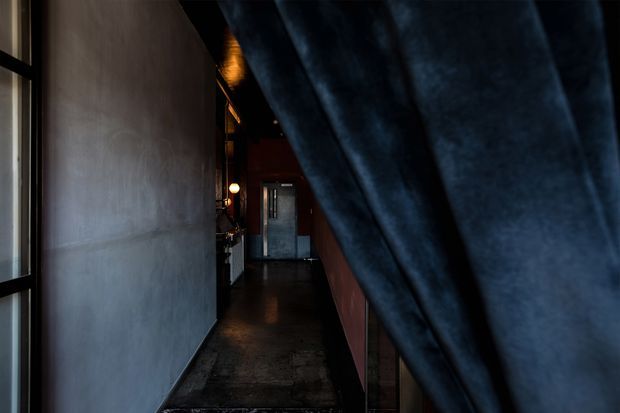 A dark, narrow interior corridor viewed beside a large dark blue draped or folded fabric element in the foreground. The hallway has a smooth grey wall, dark polished flooring, warm recessed and pendant lighting, and a doorway or glazed opening at the far end. The atmosphere is moody and industrial-leaning, with limited natural light.