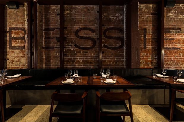 Dark, atmospheric restaurant dining area with intimate tables set with wine glasses and crockery. Exposed aged brick walls, heavy dark timber framing, black upholstered banquette seating, and warm timber dining tables create an industrial yet refined interior.