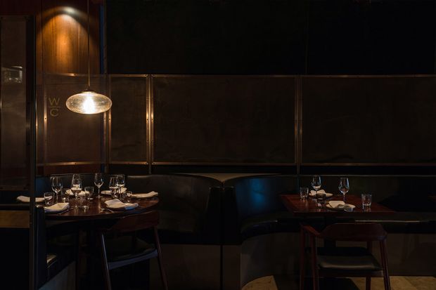 Moody, intimate restaurant dining area with dark leather banquette seating, small wood dining tables, upholstered chairs, glassware and table settings. Dark bronze or black framed wall panelling and warm pendant lighting create a refined, low-lit atmosphere.