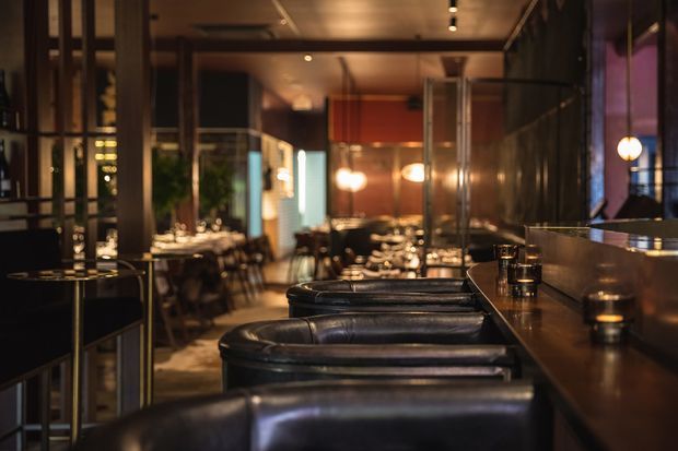 A dimly lit, sophisticated restaurant interior viewed along a long bar counter, with black leather bar stools in the foreground and dining tables extending into the background. Warm pendant and accent lighting, dark timber or metal framing, reflective surfaces, and a polished, intimate atmosphere define the space.