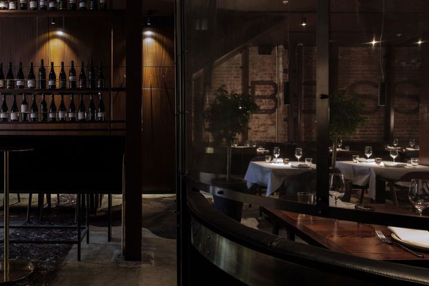 A dark, atmospheric restaurant interior with a wine display and bar area on the left, and a dining room visible through glass partitions and reflections on the right. Tables are formally set with white linens, wine glasses, plates, and cutlery. The space features exposed brick walls, dark timber or wood-toned joinery, polished concrete flooring, and dramatic warm spotlighting.
