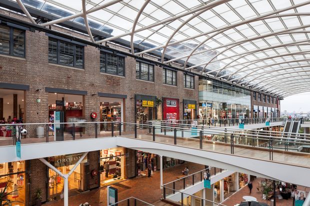 A multi-level indoor commercial shopping and retail space with exposed brick walls, rows of storefronts, pedestrian bridges, and a large translucent glazed barrel-vault roof supported by a repeating steel structure. The bright atrium includes multiple balconies, escalator or stair circulation, retail signage, and abundant overhead daylight.