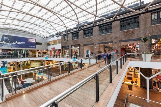 Interior view of a multi-level shopping centre with a broad timber-finished pedestrian bridge, glass and metal balustrades, brick retail frontage, numerous shopfronts, and a large translucent arched roof structure bringing in daylight. The scene is clearly a commercial retail atrium with open circulation across multiple levels.