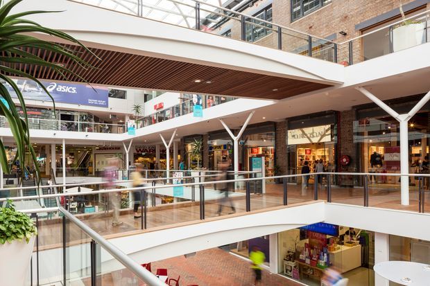 Interior view of a bright multi-level shopping centre atrium, featuring retail storefronts, open balconies with glass balustrades, white branching structural columns, exposed brick walls, and a timber slatted soffit. Shoppers and signage are visible throughout the commercial space.