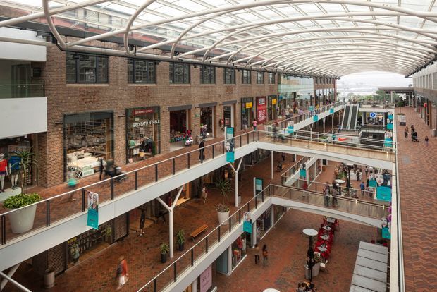A multi-level indoor shopping and commercial arcade with exposed red-brick facades, numerous retail storefronts, open balconies and walkways, escalators, and a large translucent barrel-vaulted glass canopy supported by curved metal framing. The atrium is brightly daylit and includes planted areas, seating, and pedestrian circulation across several levels.