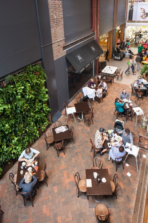 High-angle view of a lively indoor shopping-centre dining area or food-court courtyard. Numerous small dark timber tables and bentwood-style chairs are arranged on a warm reddish brick-paved floor. A tall dark charcoal facade with exposed red brick sections, a black café awning, vertical red service piping, and a large lush green living wall frame the seating area. Retail shops and pedestrians are visible in the background.