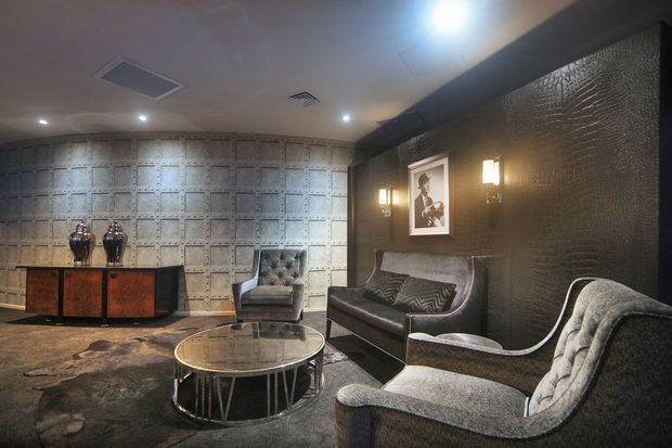 A dark, highly styled lounge or reception area with a dramatic metallic patterned feature wall, crocodile-textured dark wall covering, charcoal carpet, sculptural grey upholstered armchairs and sofa, a round glass coffee table, and a timber-and-metal sideboard. Decorative wall sconces, framed artwork, and ornate vases add a luxurious, vintage-inspired atmosphere.