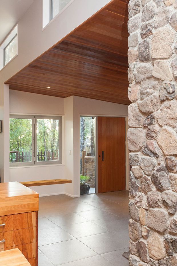 Bright contemporary residential entry area with a dramatic double-height volume, warm timber-lined ceiling, large windows, timber front door, tiled floor, built-in timber bench, and a prominent exposed rough-stone feature wall. The edge of a timber kitchen or cabinetry unit is visible in the foreground.