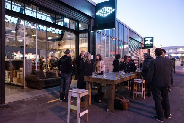 A dusk view of a contemporary commercial venue with a glazed industrial-style facade, open restaurant or bar frontage, and outdoor communal tables occupied by patrons. Dark metal framing, corrugated metal cladding, exposed structural elements, and large warehouse-like glazing define the building, while warm interior lighting creates an inviting atmosphere.