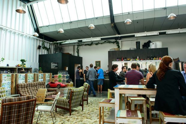 A lively industrial-style café or hospitality venue with a high glazed ceiling, exposed structural elements, pendant lighting, and a mix of communal timber tables and vintage upholstered lounge seating. A bar and drinks display occupy the rear, while greenery trails along the walls and a patterned carpet adds warmth.