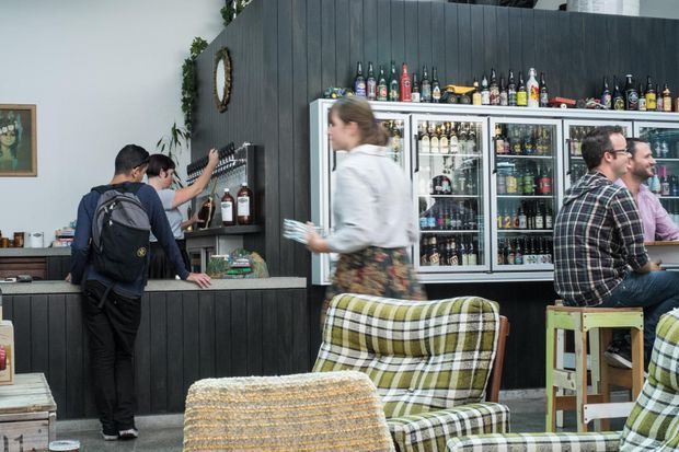 A casual bar or café interior with dark vertical wall panelling and matching counter cabinetry. A refrigerated drinks display and rows of bottles form the backdrop, while patrons sit on green-and-cream plaid upholstered seating and wooden stools. The space has an eclectic, relaxed hospitality atmosphere with vintage-inspired furniture and busy service activity.