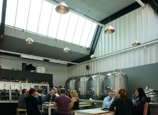 A bright industrial-style brewery taproom or hospitality space with large stainless-steel brewing tanks prominently displayed behind communal dining tables. Patrons are seated and standing around the tables, with a drinks display and service counter along the left wall. The space has corrugated metal wall cladding, exposed dark structural beams, a high sloping ceiling, extensive clerestory glazing, and multiple suspended metal pendant lights.