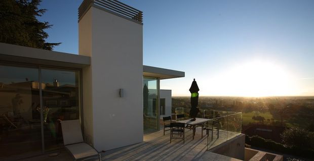 Contemporary flat-roofed residential rooftop terrace viewed at sunset, with a tall white rendered chimney or vertical architectural element capped by horizontal metal slats. Floor-to-ceiling glazing opens onto the terrace, which has a glass balustrade, outdoor dining furniture, and expansive elevated suburban views. The image is warm and backlit, with strong horizontal and vertical geometric forms.