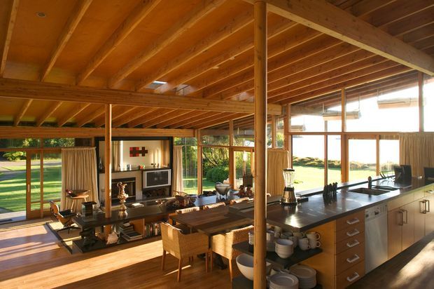 Open-plan timber-lined living, dining and kitchen interior with extensive glazing overlooking a landscaped setting. Exposed timber beams and posts form the ceiling and structure, while a dark kitchen island with light cabinetry occupies the foreground. A dining table, lounge area, fireplace or media joinery, and warm natural finishes create a spacious, sunlit retreat.