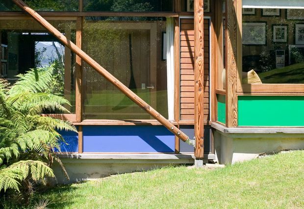 Exterior detail of a timber-framed modern dwelling, showing large glazed doors and windows, exposed diagonal timber bracing, louvred timber shutters, and brightly coloured blue and green lower wall panels. A fern-filled garden and lawn sit in front of the facade, with interior artwork and furnishings visible through the glazing.