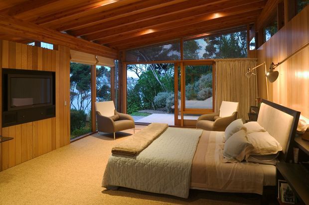 A warm, timber-lined bedroom with a pitched timber ceiling, built-in television wall, queen or king bed, bedside furnishings, and two upholstered lounge chairs. Floor-to-ceiling timber-framed glazing opens views toward a lush garden, while recessed ceiling lights and wall lamps create an intimate retreat-like atmosphere.