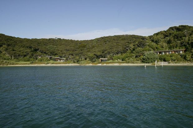 A wide exterior view across calm coastal water toward a densely forested shoreline. A few low-profile buildings are partially visible among the trees, with small waterfront structures and a narrow sandy or rocky edge along the shore. The scene is photographed in clear daylight with a natural, secluded setting.