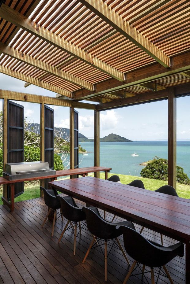 A covered outdoor dining terrace overlooking a turquoise bay, with a long dark timber dining table, black chairs, built-in barbecue, timber decking, vertical posts, and an open slatted timber pergola roof. The setting includes a lush coastal landscape, small islands, and a sailboat visible beyond the terrace.