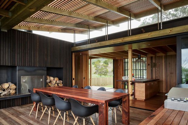 A warm, timber-lined outdoor entertaining area with a large dining table and seating, integrated fireplace, compact kitchen or bar cabinetry, and a built-in barbecue. The space is sheltered beneath a timber pergola with slatted roofing and opens onto a landscaped setting through large glazed openings.