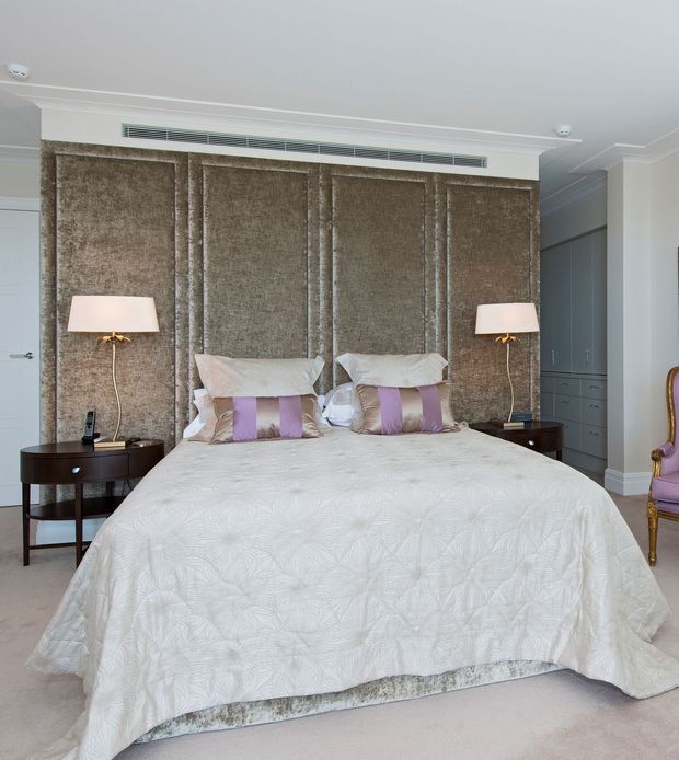 A refined bedroom with a centered upholstered bed dressed in a white patterned quilt and lavender-accented pillows. The bed is framed by a tall textured taupe feature wall with decorative panel mouldings, flanked by dark wood bedside tables and matching table lamps. Built-in grey cabinetry is visible to the right, along with a partially visible ornate purple upholstered chair. The room has pale carpet, white walls and ceiling, and a softly elegant, traditional-luxe character.