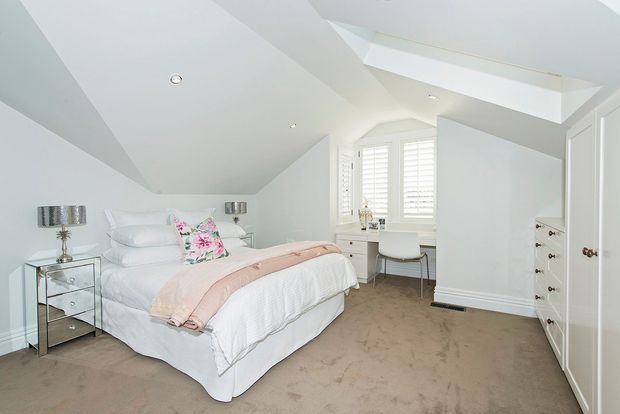 Bright, spacious bedroom with a white upholstered bed, layered white bedding, a blush pink throw, and floral accent cushions. Mirrored bedside tables and metallic lamps flank the bed. The room features white walls and built-in cabinetry, plantation shutters, a desk and chair by the window, beige carpet, and a large angled skylight that brings in abundant natural light.