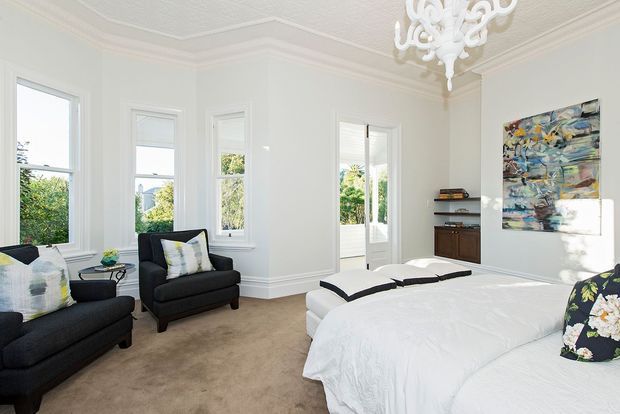 Bright, spacious bedroom with tall bay-style windows, white walls and ornate ceiling detailing, a white bed, dark upholstered seating, built-in shelving, and a large abstract artwork. Natural light creates an airy, refined atmosphere.