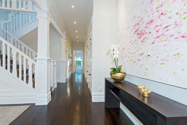 A bright, refined residential hallway and entry passage with dark timber flooring, white painted architectural trim, a substantial staircase and balustrade on the left, and a long black console beneath a large abstract artwork on the right. Recessed ceiling lights lead toward a glazed doorway at the far end, creating a strong sense of depth. The space has a polished, classic interior character with high-contrast white and dark finishes.