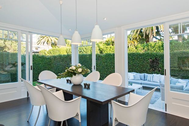 Bright dining room with a dark rectangular dining table surrounded by sculptural white chairs and a central floral arrangement. Multiple white pendant lights hang above the table. White-framed glazed doors and windows open visually onto a leafy courtyard or patio with an outdoor lounge setting. The room has dark timber flooring, white walls and trim, and a light blue ceiling, creating a fresh, airy atmosphere.