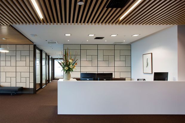 A contemporary commercial reception area with a large white reception desk, dark timber slatted ceiling, recessed linear lighting, and geometric white wall panels outlined in dark grout or trim. A floral arrangement, reception equipment, framed artwork, carpeted flooring, and glazed office partitions are visible. The space has a clean, refined, professional appearance.