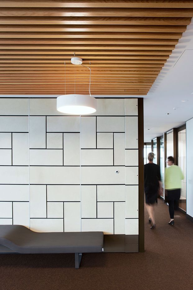 A contemporary commercial interior corridor or reception area featuring a large geometric white partition wall with dark linear joints, a sculptural white pendant light, and a built-in grey upholstered bench. The ceiling is defined by closely spaced natural timber slats, while glazed office fronts line the corridor to the right. Two people are walking through the space.