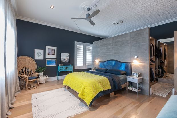 A stylish bedroom with a dark navy feature wall, timber flooring, a statement bed with a bright yellow quilt and blue upholstered headboard, woven armchair, turquoise console, framed artwork, plantation shutters, and an open walk-in wardrobe. The room has a decorative pressed-tin ceiling, ceiling fan, and warm layered lighting.