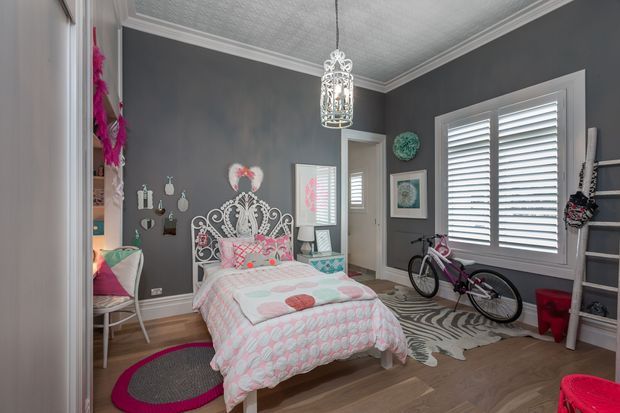A playful, brightly styled bedroom with a white ornate metal bed, pink-and-turquoise patterned bedding, charcoal grey walls, white trim, a textured white ceiling, and a decorative pendant light. Plantation shutters cover the large window, while a bicycle, zebra-pattern rug, artwork, shelving, and colourful accessories add a youthful character.
