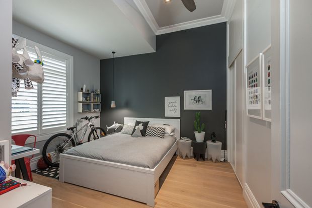 A compact, stylish bedroom with a low-profile white bed, layered grey bedding, patterned cushions, and warm timber flooring. The room has a dark charcoal feature wall with framed artwork, white built-in wardrobe doors, plantation shutters, open shelving, pendant lighting, and a bicycle stored beside the bed. A desk or study area is partially visible at the left edge.