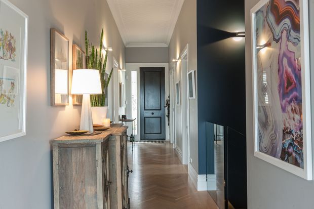 A refined residential hallway viewed toward a dark paneled entry door. The corridor features warm herringbone timber flooring, pale grey walls with white trim and cornices, framed artwork, a rustic timber console with lamp and tall indoor plants, and a dramatic dark feature wall with a linear wall light and fireplace opening. The space is long, narrow, bright, and layered with contemporary-traditional detailing.