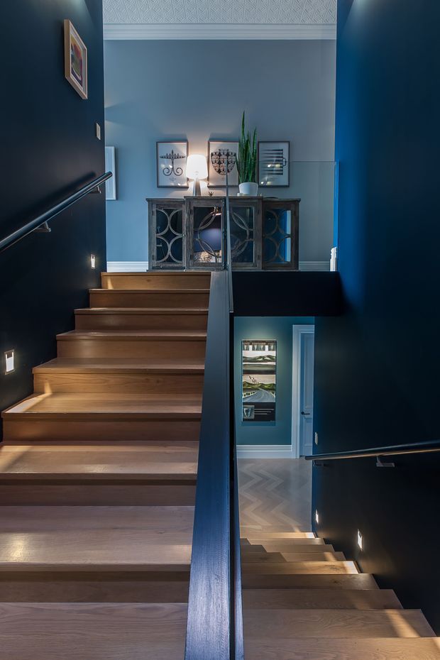 A dramatic interior view of a multi-level stairway with broad timber treads, dark navy painted walls, black handrails and balustrades, and a landing furnished with a decorative console cabinet, framed artwork, and a tall plant. A lower stair flight and adjacent room are visible through the stair opening. Decorative patterned wall panelling appears near the upper ceiling, and integrated stair lighting creates a warm, atmospheric effect.