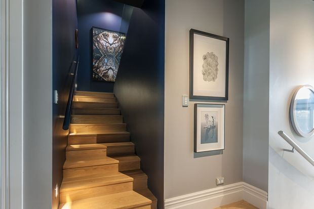 Interior view of a narrow stairway with warm timber treads and risers ascending through a dark navy-blue stairwell. Integrated step lighting highlights the staircase, while framed artwork hangs on a light grey wall beside it. A white handrail and small round window are visible on the right, with decorative wall moulding at floor level.