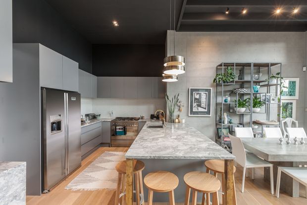 A spacious open-plan kitchen and dining area with a large marble-look island, timber stools, streamlined grey cabinetry, stainless-steel appliances, pendant lighting, and a concrete feature wall. A metal-framed open shelving unit with plants separates the kitchen from the dining area, while a partially visible dining table sits beside large windows. The space has a dark ceiling, warm timber flooring, and an industrial-contemporary aesthetic.