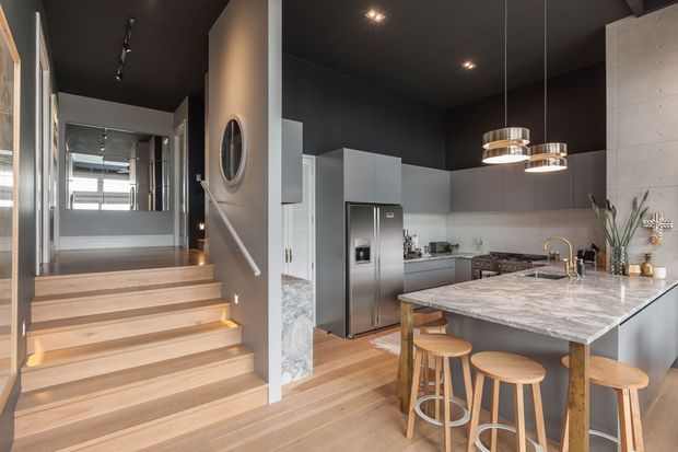 Contemporary open-plan kitchen viewed from a stepped entry or living area. The space features a large marble-look island with seating, pale grey cabinetry, stainless-steel appliances, light timber flooring and stairs, a dark ceiling, pendant lighting, and a textured light wall surface. A mirrored wall and hallway are visible beyond the stairs.