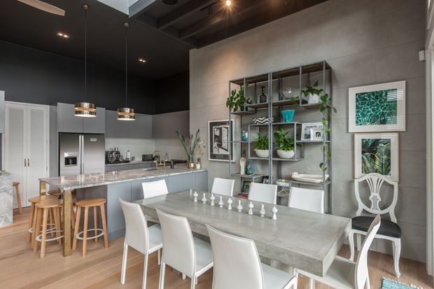 Open-plan contemporary kitchen and dining area with a large pale stone dining table, white upholstered chairs, a stone kitchen island with timber stools, grey cabinetry, stainless-steel appliances, and dark ceilings. A black-framed open display shelf with plants and decorative objects sits against a concrete-look feature wall, creating an industrial-modern character.