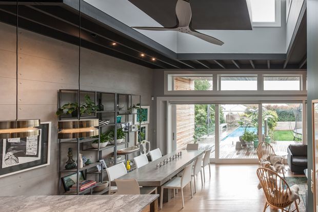 Open-plan dining and living area with a long dining table, contemporary shelving, pendant lights, and large glazed doors opening to a deck and pool area. The room features a concrete-look wall, timber flooring, a marble-look kitchen benchtop in the foreground, and a large ceiling fan beneath a skylight or clerestory opening.