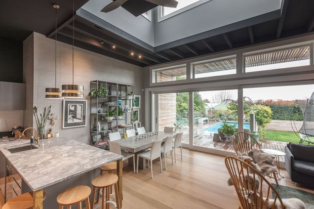 Open-plan kitchen and dining area with a large marble-look island and timber stools, pendant lighting, exposed dark ceiling beams, a skylight, built-in shelving, and extensive floor-to-ceiling glazing. The dining area opens visually to an outdoor pool and garden, while a small living area is visible at right. The space has a warm contemporary character with strong indoor-outdoor connection and abundant natural light.