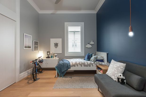 A compact, carefully styled bedroom with blue-grey walls, white trim and built-in wardrobe doors. The room contains a single bed with patterned bedding, a grey upholstered sofa, a white dresser, bicycle storage, framed wall art, a window with white plantation shutters, a pendant light and light timber flooring. The space has a youthful, practical feel with a small rug and soft neutral furnishings.