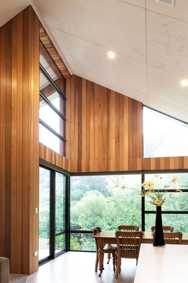A bright double-height dining area with extensive floor-to-ceiling glazing overlooking dense greenery. Vertical warm timber wall panelling wraps the room, contrasting with a pale timber-lined ceiling and dark-framed windows and doors. A timber dining table and chairs sit beside a white kitchen island or counter, with a black vase and flowers as a small focal point.