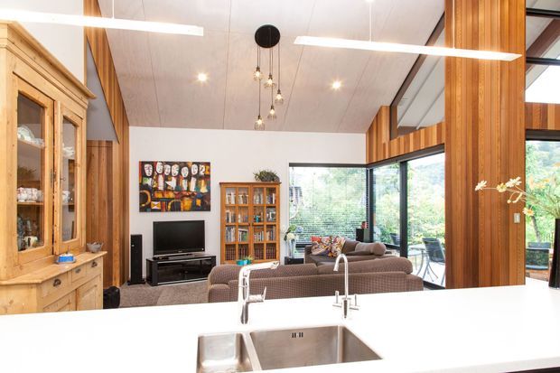 Open-plan kitchen and living area with a bright white island and stainless-steel double sink in the foreground. The living room includes a sectional sofa, television, timber display cabinetry, and a large timber-framed glazed corner opening to greenery outside. The space has a high sloped timber-lined ceiling, exposed timber framing and cabinetry, recessed lighting, and suspended linear and pendant lights.