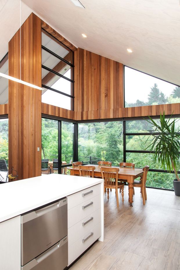 Bright open-plan kitchen and dining area with a white island featuring integrated stainless-steel appliances and drawers. The room has extensive floor-to-ceiling black-framed glazing overlooking lush greenery, a warm vertical timber-lined feature wall, light timber flooring, a pitched ceiling with skylights, and a traditional timber dining suite. A large indoor plant is visible near the glazing.